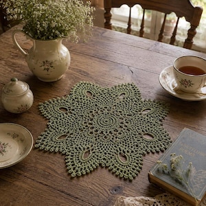 May include: A green crocheted doily with a starburst design sits on a wooden table. A floral teacup and saucer, a small sugar bowl, and a vase of flowers are also on the table. A vintage book is on the right side of the doily.