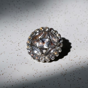 May include: A silver-toned brooch with a round, faceted crystal center surrounded by smaller crystals. The brooch has a starburst design.