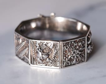 May include: A silver-toned hinged bracelet with an octagonal design. Each panel features detailed floral and botanical relief carvings, including roses, wheat, and other foliage. The bracelet is closed with a clasp.