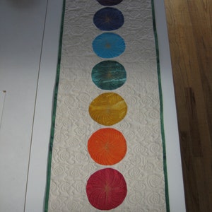 May include: A white table runner with a green border and seven embroidered circles in a rainbow color pattern. The circles are arranged vertically in a line down the center of the runner.