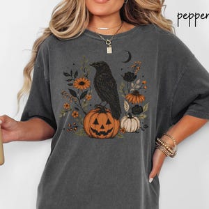 May include: A charcoal gray short-sleeve t-shirt featuring a graphic print of a black crow perched on a carved jack-o'-lantern pumpkin, surrounded by orange flowers, dark foliage, and a crescent moon. The word pepper is printed in the top right corner.