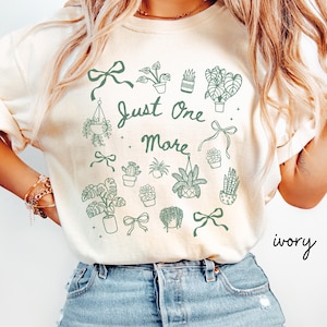 K&ouml;nnte beinhalten: Elfenbeinfarbenes Sweatshirt mit gr&uuml;nem botanischem Design. Das Design umfasst verschiedene Topfpflanzen, Schleifen und den Text "Just One More". Das Wort "ivory" ist rechts unten in Kursivschrift geschrieben.