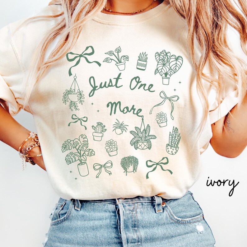 K&ouml;nnte beinhalten: Elfenbeinfarbenes Sweatshirt mit gr&uuml;nem botanischem Design. Das Design umfasst verschiedene Topfpflanzen, Schleifen und den Text "Just One More". Das Wort "ivory" ist rechts unten in Kursivschrift geschrieben.