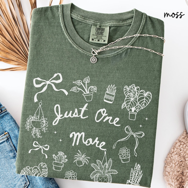 K&ouml;nnte beinhalten: Moosgr&uuml;nes T-Shirt mit wei&szlig;er Grafik von Topfpflanzen und dem Text "Just One More". Eine silberne Halskette mit Anh&auml;nger liegt auf dem Shirt. Das Shirt ist auf einer wei&szlig;en Oberfl&auml;che mit einer Jeans gefaltet.
