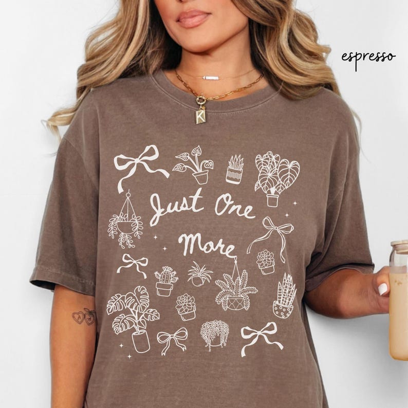 K&ouml;nnte beinhalten: Espressofarbenes T-Shirt mit dem Text "Just One More" und wei&szlig;en Illustrationen von Pflanzen und Schleifen. Das Shirt hat einen Rundhalsausschnitt und kurze &Auml;rmel. Das Wort "espresso" steht in Kursivschrift oben rechts.
