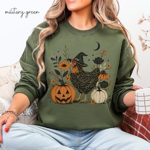 May include: A military green crewneck sweatshirt featuring a graphic print of a chicken wearing a witch hat, surrounded by sunflowers, foliage, a crescent moon, and two pumpkins, one of which is a jack-o'-lantern with a carved face.