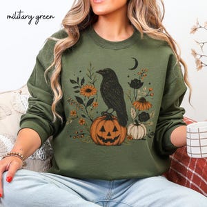 May include: A military green crewneck sweatshirt featuring a graphic print of a black crow perched on a carved jack-o'-lantern pumpkin. The design includes orange and black floral elements, berries, and a crescent moon, all centered on the chest.
