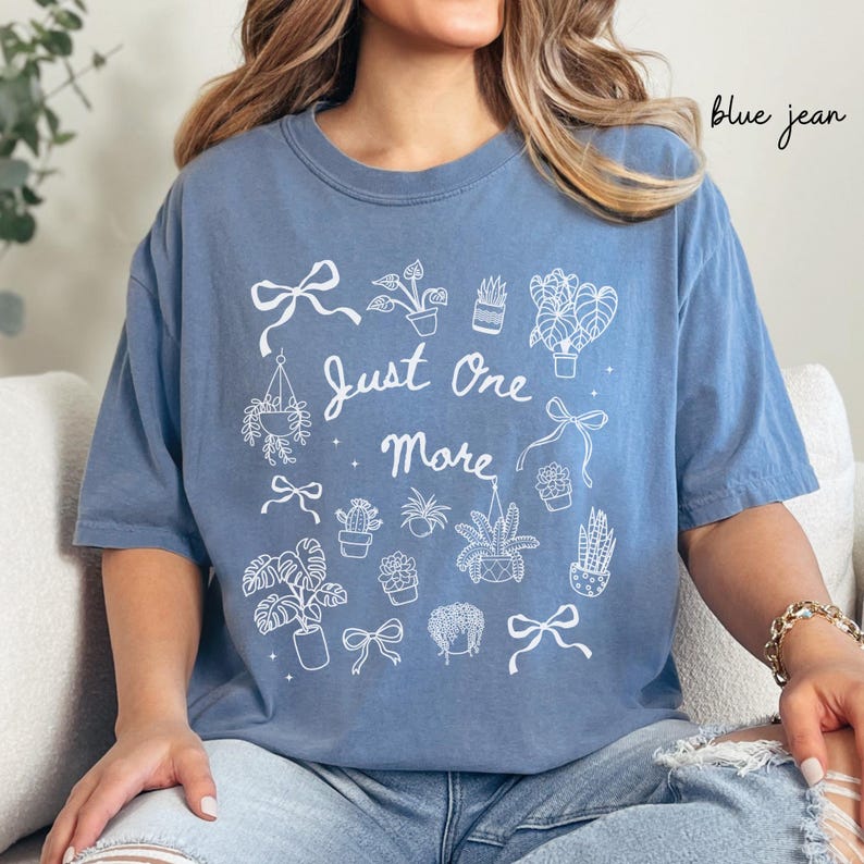 K&ouml;nnte beinhalten: Blaues Jeans-T-Shirt mit dem Text "Just One More" und wei&szlig;en Illustrationen von Pflanzen und Schleifen. Das Shirt hat einen l&auml;ssigen Stil.