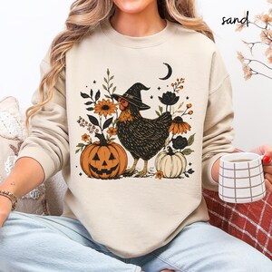 May include: A sand-colored crewneck sweatshirt featuring a graphic print of a chicken wearing a witch hat, surrounded by orange and black flowers, a crescent moon, and two pumpkins, one orange and one white, with the word sand printed above.