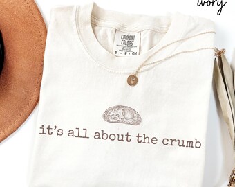 May include: Ivory-colored t-shirt with the phrase "it's all about the crumb" printed in brown. The shirt features a graphic of a bread crumb and a gold necklace with a pendant. A brown hat is visible in the upper left corner.