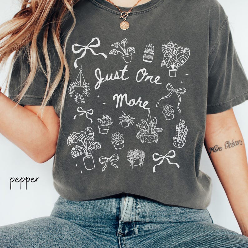 K&ouml;nnte beinhalten: Dunkelgraues T-Shirt mit einer wei&szlig;en Grafik von Topfpflanzen, Schleifen und dem Text "Just One More". Das Shirt besteht aus einem weichen Material und hat eine lockere Passform. Das Wort "pepper" ist unten links aufgedruckt.