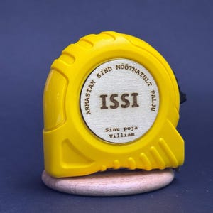 Puede incluir: Un flex&oacute;metro amarillo con la palabra "ISSI" en el centro de un c&iacute;rculo de madera. El texto "Armastan sind m&otilde;&otilde;tmatult palju" rodea el c&iacute;rculo. El texto "Sinu poja Villiam" est&aacute; debajo del c&iacute;rculo.