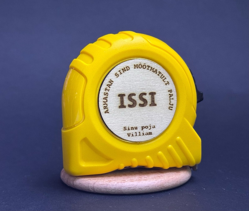 Puede incluir: Un flex&oacute;metro amarillo con la palabra "ISSI" en el centro de un c&iacute;rculo de madera. El texto "Armastan sind m&otilde;&otilde;tmatult palju" rodea el c&iacute;rculo. El texto "Sinu poja Villiam" est&aacute; debajo del c&iacute;rculo.