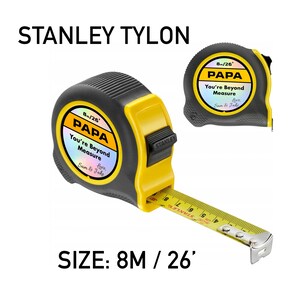 Puede incluir: Un flex&oacute;metro Stanley Tylon negro y amarillo con el texto "PAPA You're Beyond Measure Love Sam & Jake". El flex&oacute;metro mide 8 metros (26 pies). El texto "STANLEY TYLON" est&aacute; en la parte superior.