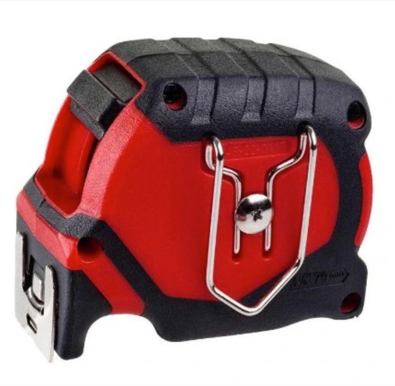 Puede incluir: Un flex&oacute;metro rojo y negro con un clip met&aacute;lico para cintur&oacute;n. El flex&oacute;metro tiene una parte superior negra texturizada y un cuerpo rojo. La cinta se extiende desde la parte inferior y tiene medidas en cm y metros.
