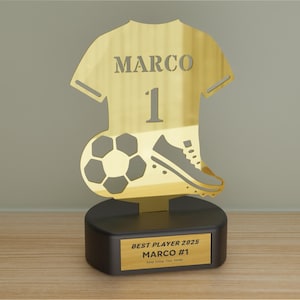 Può includere: Un trofeo di calcio dorato con la sagoma di una maglia con il nome "MARCO" e il numero "1". Sotto la maglia ci sono un pallone da calcio e una scarpa da calcio. La base nera ha una placca dorata con la scritta "BEST PLAYER 2025 MARCO #1".