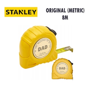 Puede incluir: Un flex&oacute;metro Stanley amarillo de 8 m con el texto "DAD" y "No One Measures Up To You". El flex&oacute;metro est&aacute; extendido, mostrando las marcas de medici&oacute;n. El logotipo de Stanley es amarillo.