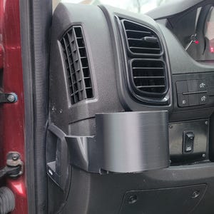 May include: A black plastic cup holder is mounted on the side of a car's dashboard. The cup holder is located near the driver's side door and has a black plastic vent above it.