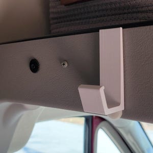 May include: White plastic hook mounted on a gray surface. The hook is designed to hang items from the ceiling of a vehicle.
