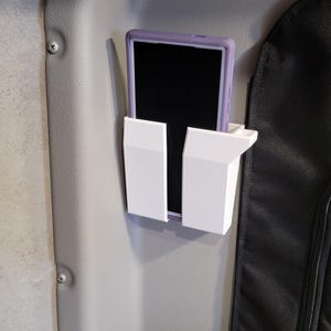 May include: A white plastic phone holder mounted to a gray surface. The holder has two sections, one on each side of the phone, to keep it secure. A purple phone case is visible in the holder.