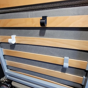 May include: Close-up of a wooden slat bed frame with three white plastic hooks and one black plastic hook attached to the slats.