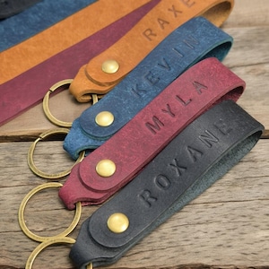 Peut inclure: Plusieurs porte-clés personnalisés en cuir de différentes couleurs, dont noir, bordeaux, bleu et beige. Chaque porte-clés porte un nom gaufré en majuscules, fixé avec un anneau en métal couleur laiton et une fermeture à bouton-pression.