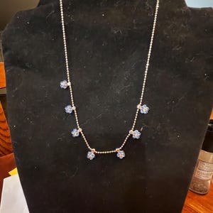 May include: A silver-toned necklace with small, light blue flower pendants. The necklace is displayed on a black velvet bust. The flowers have a delicate, detailed design, and the chain is made of small, round beads.