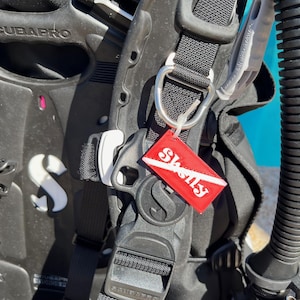 Puede incluir: Un BCD de buceo negro con una etiqueta roja "Shelly" unida a un anillo en D. El BCD tiene una correa negra con una hebilla plateada y una botella de buceo negra.