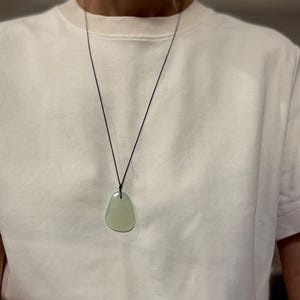 May include: A light green, irregularly shaped pendant necklace. The pendant hangs from a thin, black cord. The necklace is worn over a white t-shirt.