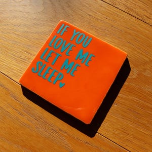 Könnte beinhalten: Oranges Quadrat mit dem Text "IF YOU LOVE ME LET ME SLEEP" in Türkis. Das Quadrat liegt auf einer Holzoberfläche. Der Text ist in einer serifenlosen Schriftart.