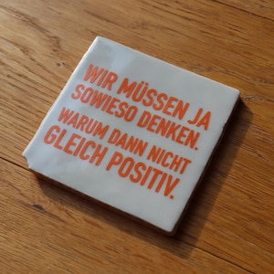 Könnte beinhalten: Ein quadratischer, cremefarbener Untersetzer mit orangefarbenem Text. Der Text lautet: "WIR MÜSSEN JA SOWIESO DENKEN. WARUM DANN NICHT GLEICH POSITIV." Der Untersetzer liegt auf einer Holzoberfläche.