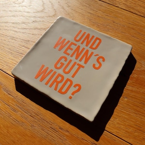 Könnte beinhalten: Ein quadratischer, hellgrauer Keramikuntersetzer mit dem deutschen Text "UND WENN'S GUT WIRD?" in Orange. Der Untersetzer hat einen leicht unebenen Rand und liegt auf einer Holzoberfläche.