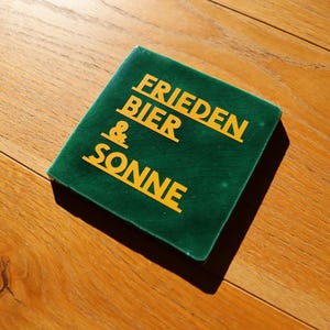 Könnte beinhalten: Ein quadratischer, grüner Untersetzer mit den Worten "FRIEDEN BIER & SONNE" in gelben Buchstaben. Der Untersetzer liegt auf einer Holzoberfläche. Der Text ist in einer fetten, serifenlosen Schriftart.