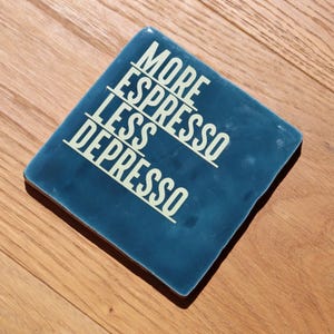 Könnte beinhalten: Ein quadratischer, tiefblauer Keramikuntersetzer mit glänzender Oberfläche. Der Text "MORE ESPRESSO LESS DEPRESSO" ist in einer fetten, cremeweißen serifenlosen Schrift mittig aufgedruckt, wobei horizontale Linien die drei Textzeilen trennen.