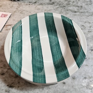 May include: A round ceramic bowl featuring a white base with vertical, hand-painted dark green stripes. The bowl has a smooth, glossy finish and a wide, circular opening, showcasing a consistent pattern of alternating green and white bands.