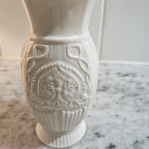 May include: A white ceramic vase with a detailed embossed design. The vase features a flared top, a detailed central section with an ornate pattern, and a pedestal base. The vase is approximately 8 inches tall.