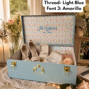 May include: A light blue trunk with gold-tone latches and handle, featuring an interior floral pattern. The lid interior displays the embroidered text "The Weiler's est. 2026". Inside are white high-heeled sandals, a ring box, and a cloth with "H & J".
