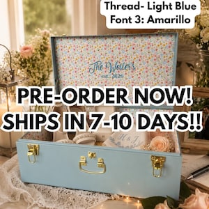 May include: A light blue storage trunk with gold-tone latches and a handle. The interior lid features a floral pattern and the embroidered text "The Weiler's est. 2026". Inside are white shoes, a small box, and a cloth with "H & J" embroidery.