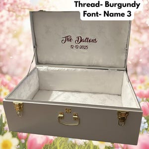 May include: A white trunk with gold-colored hardware, open to reveal a white interior with a floral pattern. The text "The Daltons" and the date "12-12-2025" are embroidered in burgundy thread on the inside of the lid. The trunk is set against a floral background.