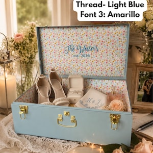 May include: A large light blue trunk with gold-tone latches and handle. The interior is lined with a floral pattern and features the embroidered text "The Weilers est. 2026". Inside are white high-heeled sandals, a ring box, and a cloth with "H & J".