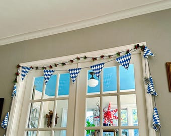 Op de afbeelding: Een feestelijke slinger van blauw-witte ruitvormige vlaggetjes hangt boven een witomrande glazen deur. De slinger is versierd met kleine rode en groene decoratieve elementen, wat een vrolijke, feestelijke sfeer creëert.