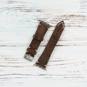 May include: Two-piece brown leather watch band with white stitching. The band has a silver buckle and is designed to fit a smartwatch. The leather has a distressed look, and the band is lying on a white wooden surface.