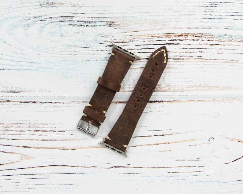 May include: Two-piece brown leather watch band with white stitching. The band has a silver buckle and is designed to fit a smartwatch. The leather has a distressed look, and the band is lying on a white wooden surface.