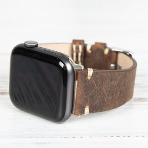 May include: A black smartwatch with a brown leather band. The band has a distressed texture and cream-colored stitching. The watch is displayed on a white surface.