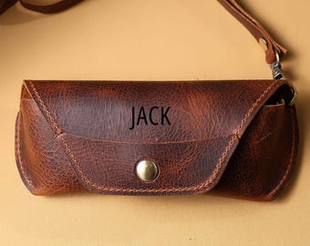 May include: A brown leather glasses case with the name "JACK" printed on the front. The case has a snap closure and a wrist strap. The leather has a distressed look.