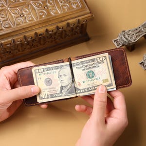 May include: A brown leather money clip holding two ten-dollar bills. The bills display the text "THE UNITED STATES OF AMERICA" and the number "10". The money clip is open, revealing the bills.
