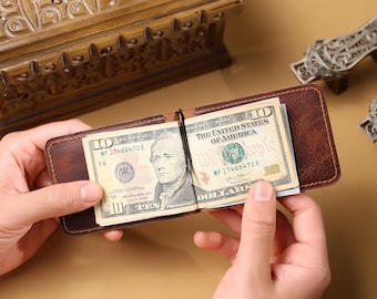 May include: A brown leather money clip holding two ten-dollar bills. The bills display the text "THE UNITED STATES OF AMERICA" and the number "10". The money clip is open, revealing the bills.