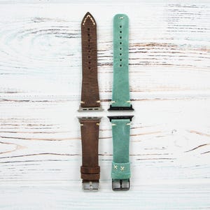 May include: Two leather watch bands, one brown and one teal, are displayed against a white wood grain background. The brown band has white stitching and a silver buckle. The teal band has a cross-stitch design and a silver buckle. Both bands are for a smartwatch.