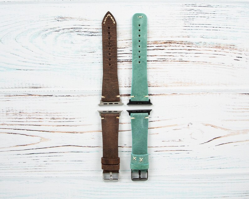 May include: Two leather watch bands, one brown and one teal, are displayed against a white wood grain background. The brown band has white stitching and a silver buckle. The teal band has a cross-stitch design and a silver buckle. Both bands are for a smartwatch.