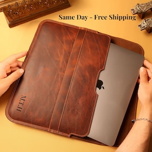 May include: A brown leather laptop sleeve with a smooth, distressed texture and rounded corners. The sleeve features a vertical strap detail and the embossed initials M.T.H. on the lower left side. A silver laptop is partially inserted into the sleeve.